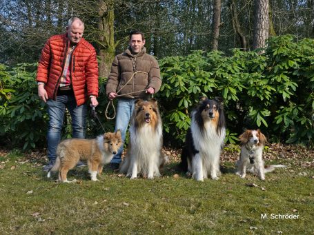 Zwei Männer stehen mit vier Hunden in einer grünen Umgebung.