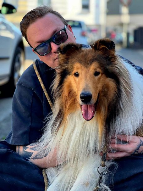 Mann mit Sonnenbrille sitzt neben einem lächelnden Collie-Hund.