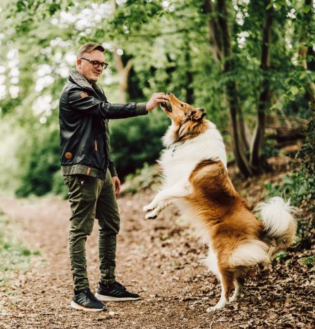 Ein Mann in einer Lederjacke streichelt einen aufrecht stehenden Collie im Wald.